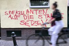 Eine Fahrradfahrerin fährt an einer Wand mit dem Graffiti "Spekulanten auf die Straße setzen" vorbei.